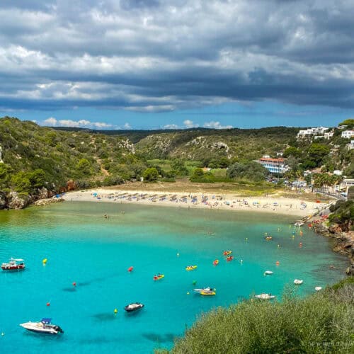 Cala en Turqueta in Menorca, Spain - a beautiful sandy beach framed by rocky cliffs and pine-covered hills, with turquoise waters dotted with boats and kayaks under a dramatic sky.