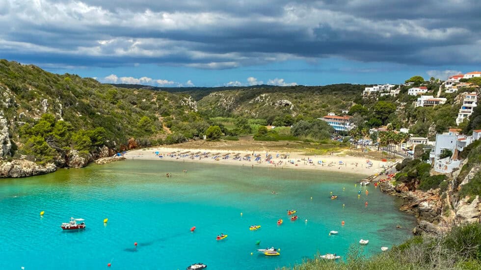 Cala en Turqueta in Menorca, Spain - a beautiful sandy beach framed by rocky cliffs and pine-covered hills, with turquoise waters dotted with boats and kayaks under a dramatic sky.