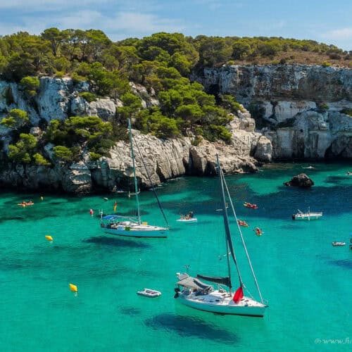 Cala Macarella in Menorca, Spain. A stunning turquoise cove surrounded by white limestone cliffs and pine trees, with sailboats and kayakers enjoying the crystal-clear Mediterranean water.