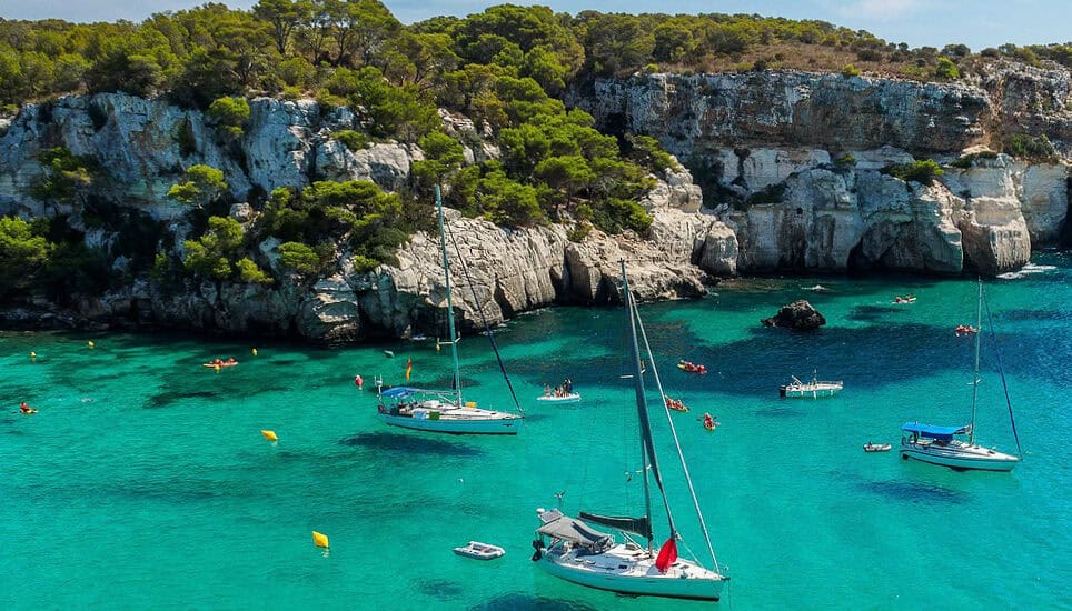 Cala Macarella in Menorca, Spain. A stunning turquoise cove surrounded by white limestone cliffs and pine trees, with sailboats and kayakers enjoying the crystal-clear Mediterranean water.