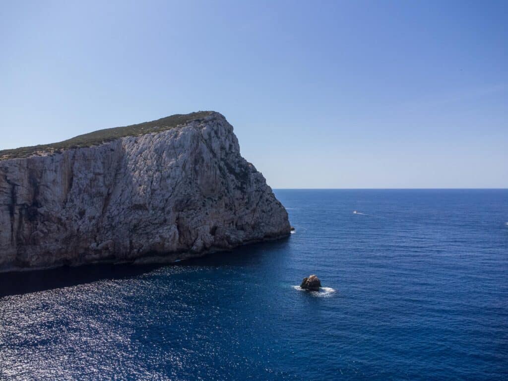 Visiting the limestone cliffs of Capo Caccia as part of a Northern Sardinia itinerary. Cliffs rising out of the blue sea, with a light blue sky.