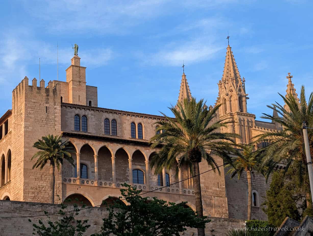 ChatGPT said: Exterior view of the Royal Palace of La Almudaina in Palma de Mallorca, showing its fortified stone walls, crenellated towers, arched windows and adjacent Gothic architectural elements beneath a blue sky.