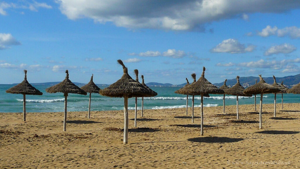 Beach with straw umbrellas and sandy shore under blue sky and clouds, scenic coastal landscape with distant mountains, relaxation spot on sunny seaside day.