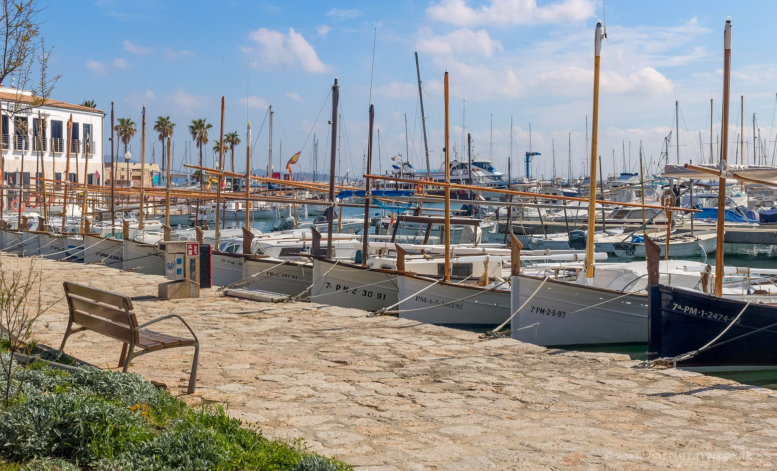 Harbour scene in Port de Pollença, Mallorca, showing moored boats bobbing on calm blue water, lined by a marina promenade and backed by distant hills under a clear sky.