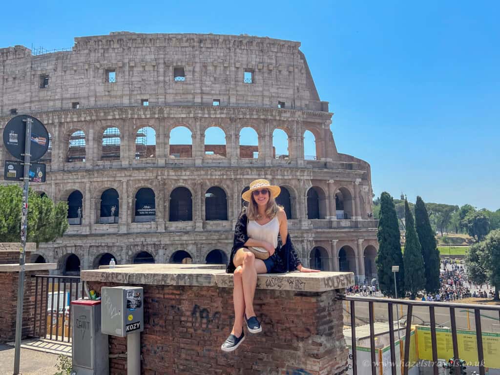 Woman sitting on a brick ledge smiling in front of the Colosseum in Rome, Italy, wearing a sunhat, sunglasses, and summer outfit on a bright sunny day.