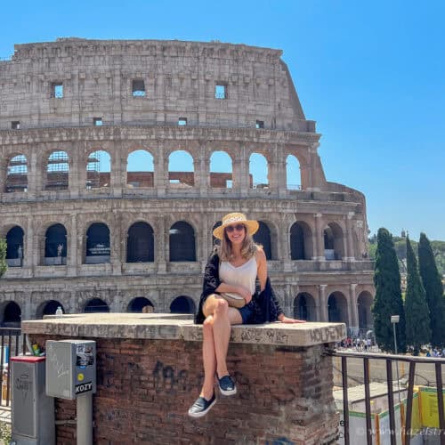 Woman sitting on a brick ledge smiling in front of the Colosseum in Rome, Italy, wearing a sunhat, sunglasses, and summer outfit on a bright sunny day.