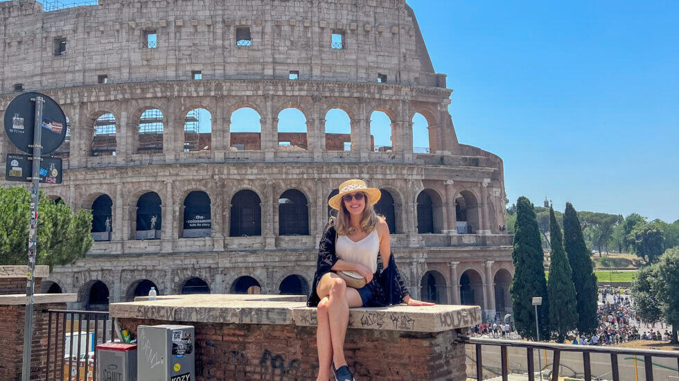 Woman sitting on a brick ledge smiling in front of the Colosseum in Rome, Italy, wearing a sunhat, sunglasses, and summer outfit on a bright sunny day.