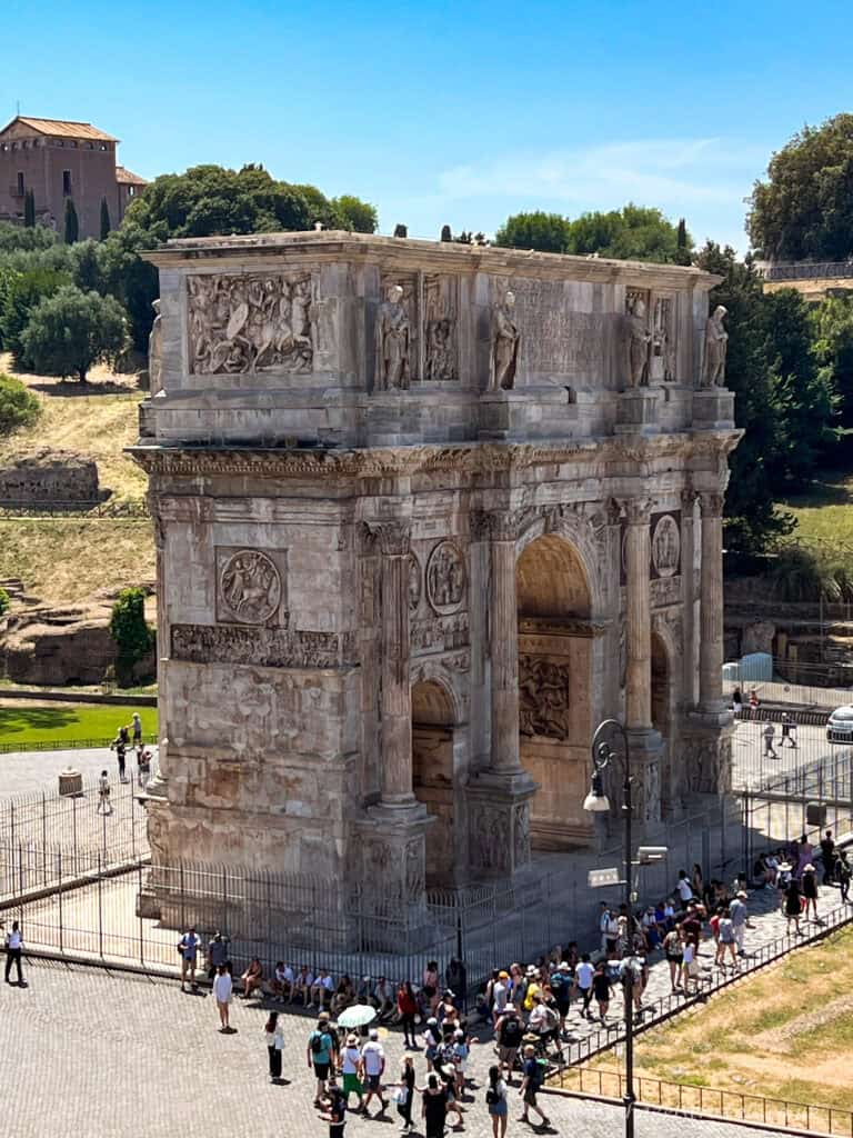 Aerial view of the Arch of Constantine in Rome, Italy, with tourists gathered around the ancient marble monument on a sunny day.