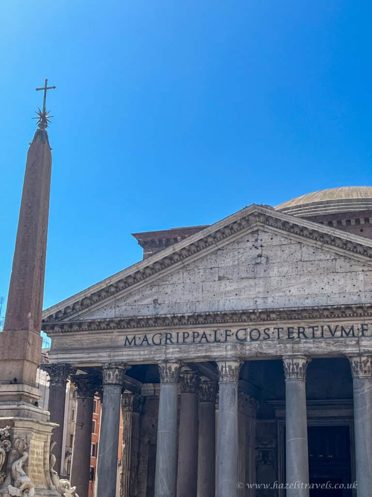 Close-up view of the Pantheon in Rome, Italy, showing its ancient stone columns, Latin inscription, and the nearby obelisk under a clear blue sky.
