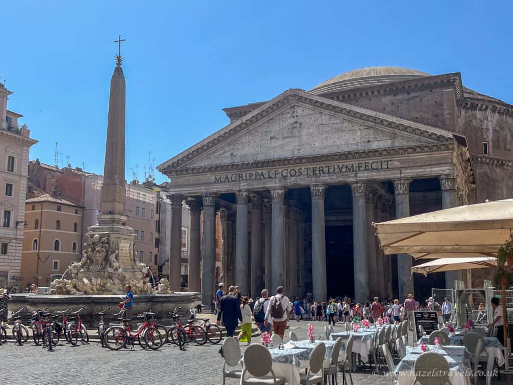 View of the Pantheon in Rome, Italy, with its grand columns and dome, lively crowds in Piazza della Rotonda, café tables in the foreground, and the Fontana del Pantheon with an obelisk under a bright blue sky.
