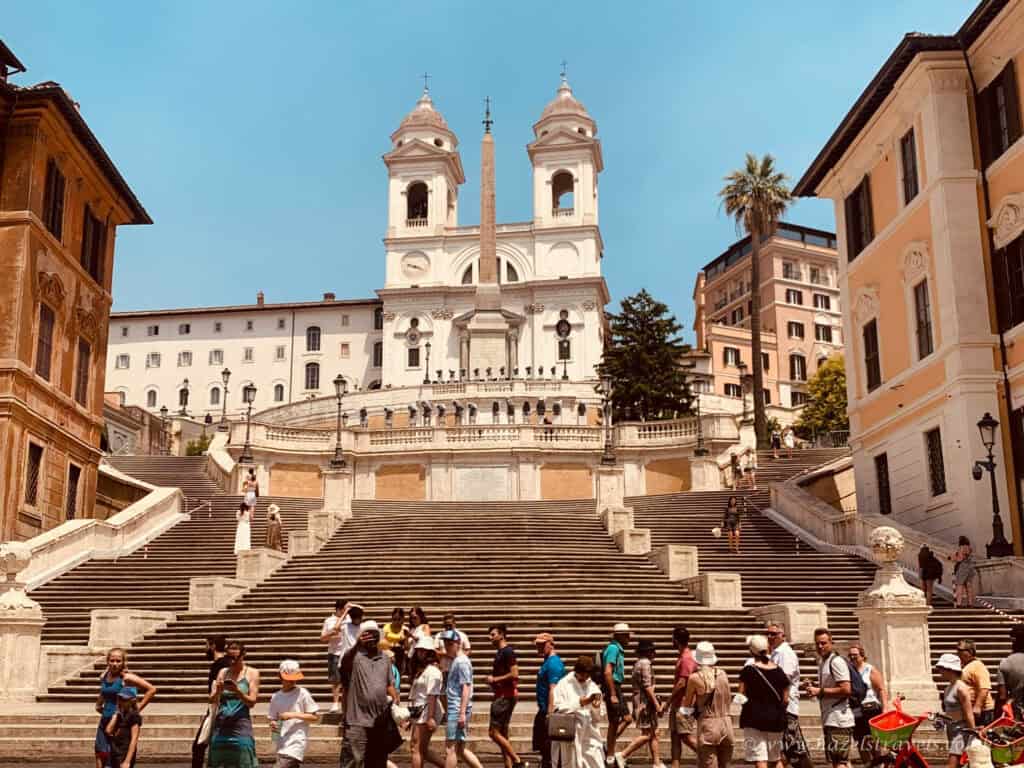 Tourists walking and relaxing on the Spanish Steps in Rome, Italy, with the Trinità dei Monti church and obelisk at the top under a bright summer sky.