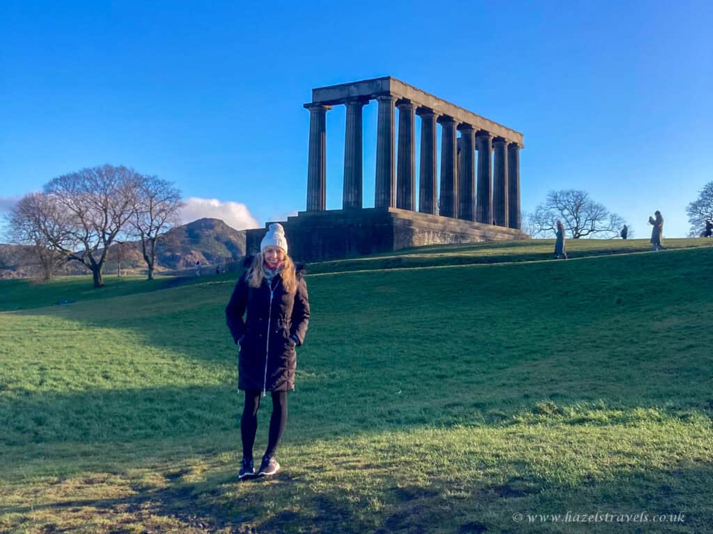 Person walking on Calton Hill, Edinburgh, with the National Monument in the background under a clear blue sky.