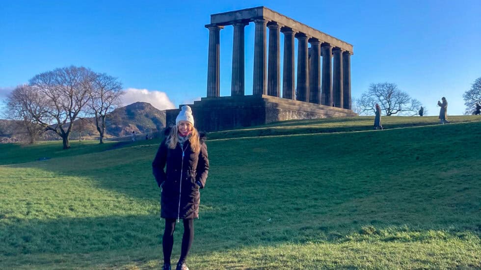 Person walking on Calton Hill, Edinburgh, with the National Monument in the background under a clear blue sky.
