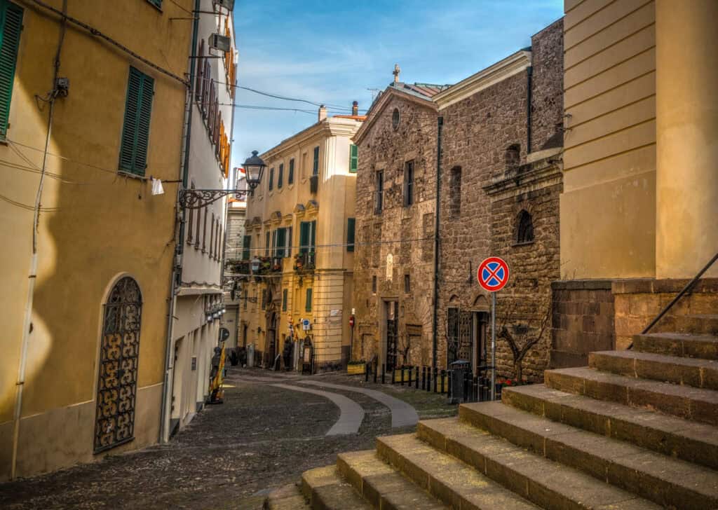Old town of Algheoa, Sardinia. Narrow cobbled street lined with pastel houses and a historic church façade in the background.
