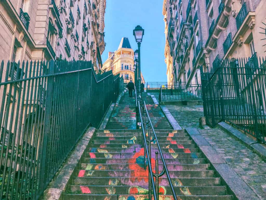 Colourful painted staircase in Montmartre, Paris, leading uphill between classic Haussmann-style buildings under a bright blue sky.