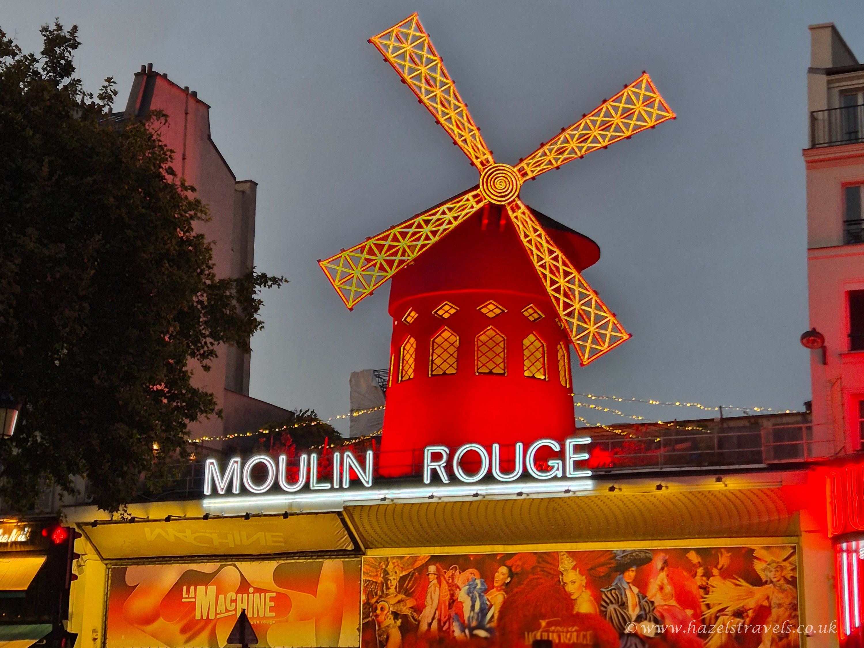 The illuminated red windmill of the Moulin Rouge cabaret in Paris at dusk, with neon lights and colourful posters below.