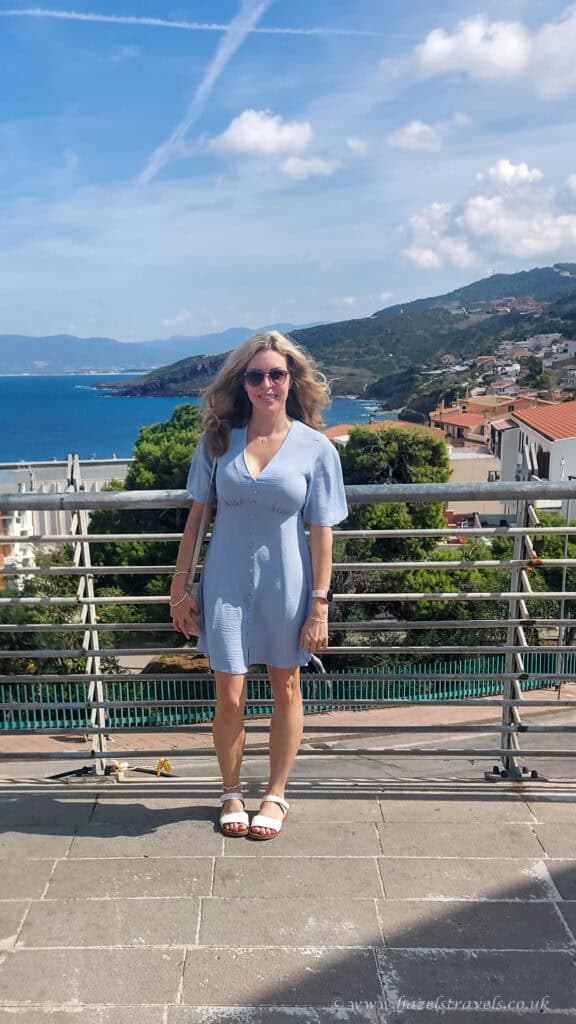 Castelsardo, Sardinia - Woman standing on a bridge overlooking the colourful coastal town of Castelsardo with hills and sea in the background.