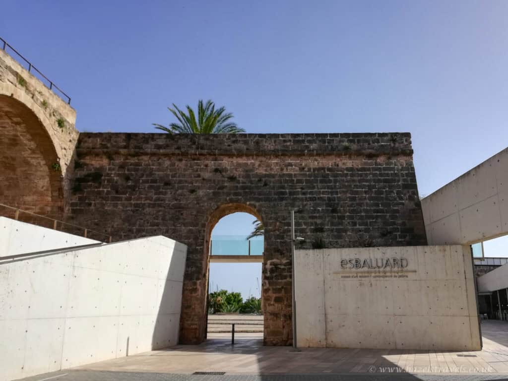 Ancient stone fortress wall with modern entrance at Esbalurd, Spain, under a clear blue sky, featuring a historic archway blend with contemporary architecture.