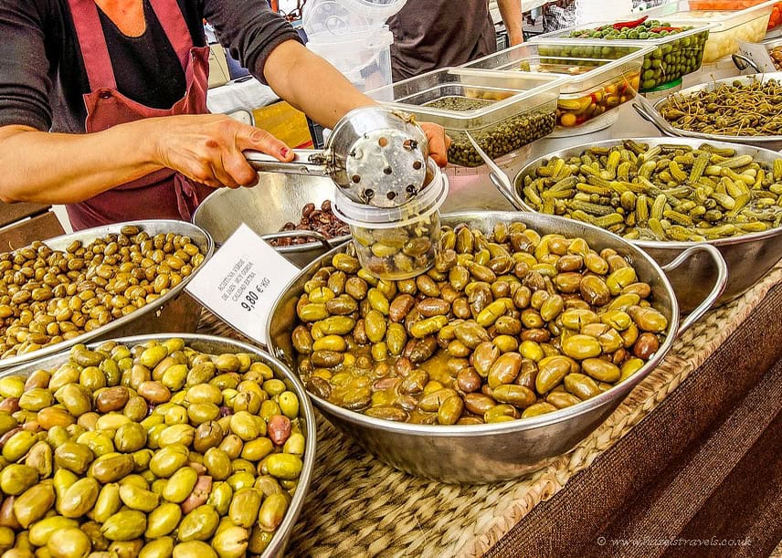 Market stall with large metal bowls filled with assorted green and brown olives, pickles, and capers, as a vendor scoops olives into a plastic container using a slotted spoon in Palma de Mallorca.