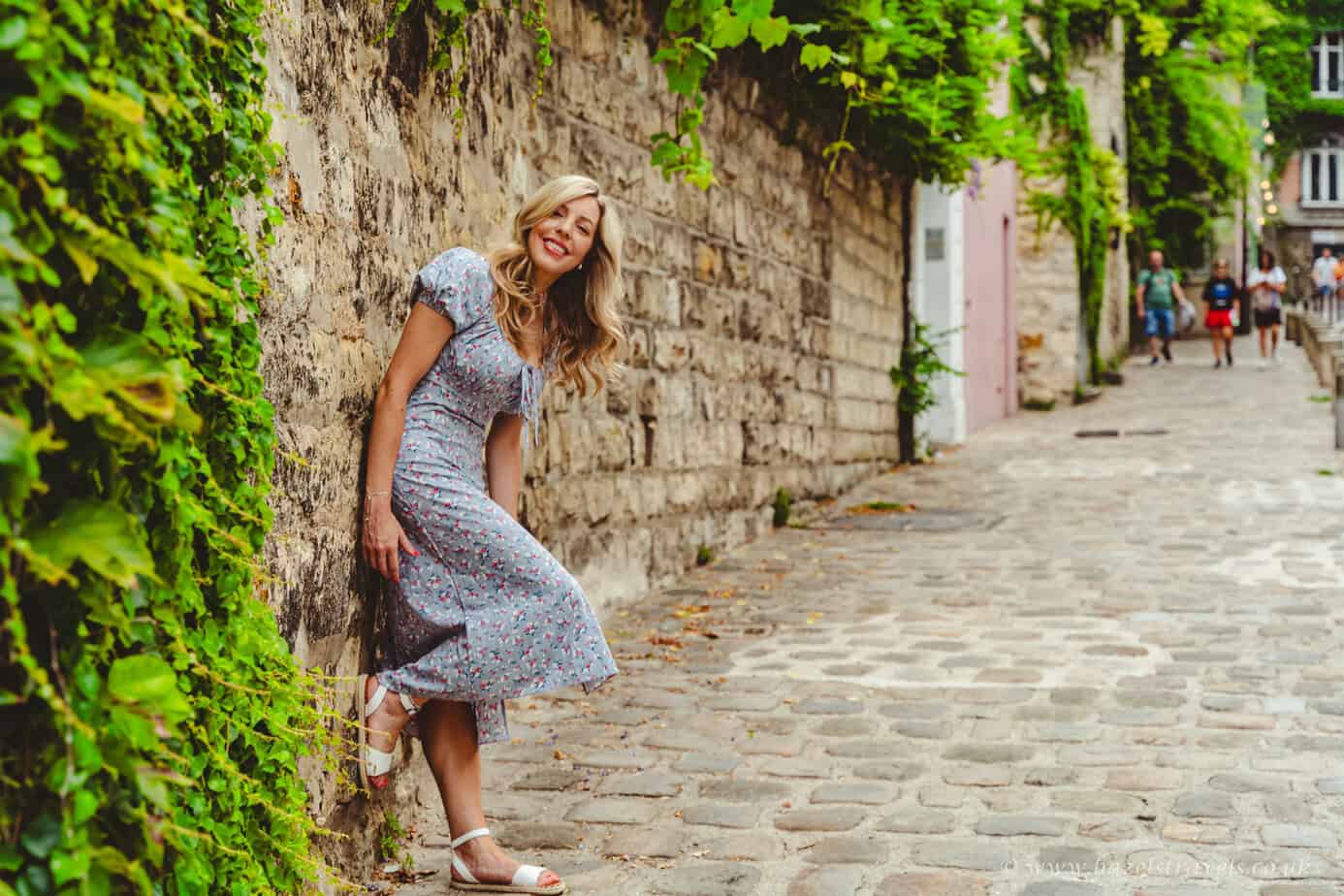 Woman in a blue patterned dress smiling while walking up a cobbled lane lined with ivy-covered stone walls in Paris.