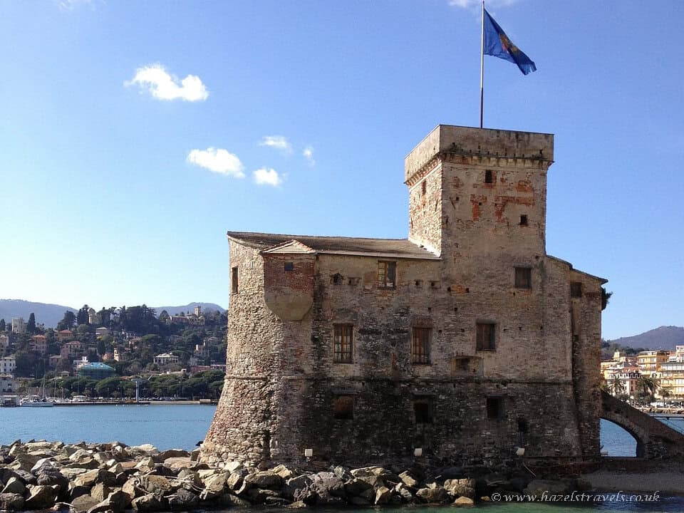 Historic seaside castle in Rapallo, Italy, known as Castello sul Mare, standing on the rocky shoreline with a flag flying above and pastel coastal buildings in the background.