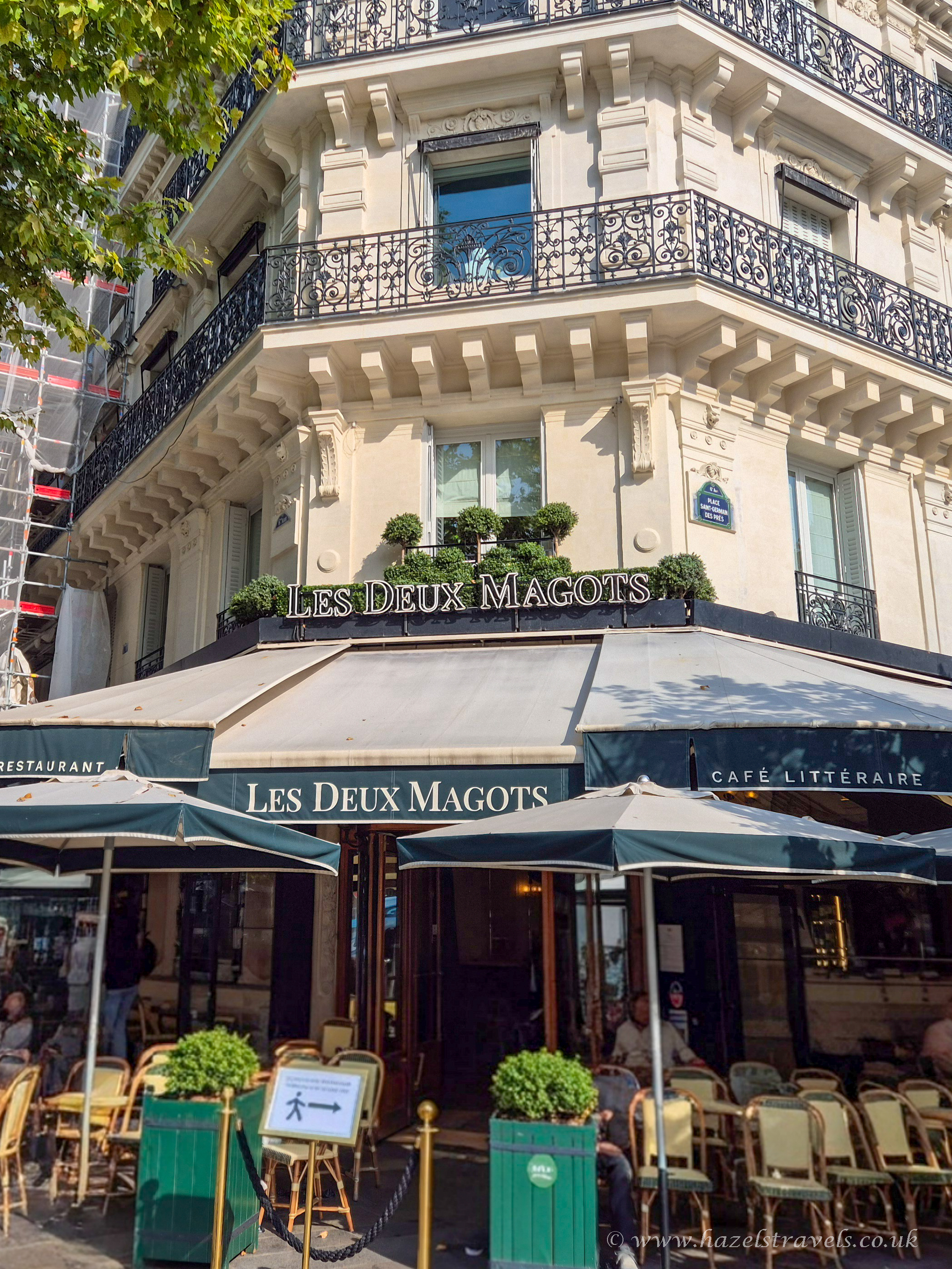 The famous Les Deux Magots café in Saint-Germain-des-Prés, Paris, with outdoor seating, green umbrellas, and classic Parisian architecture.