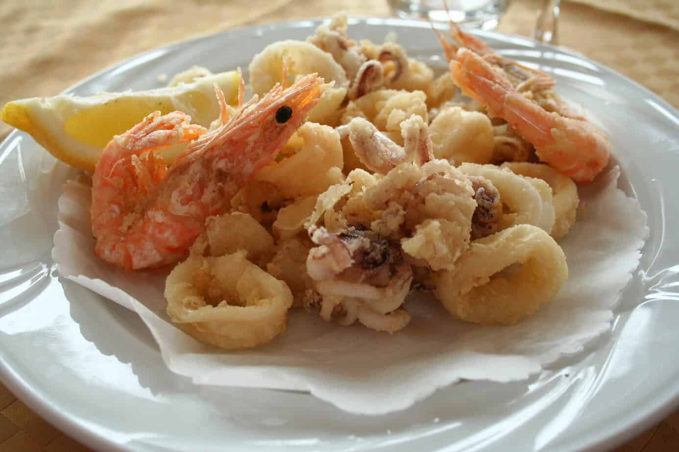Close-up of a plate of Ligurian fried seafood, featuring prawns, calamari rings, and small squid served with a wedge of lemon on a white dish.
