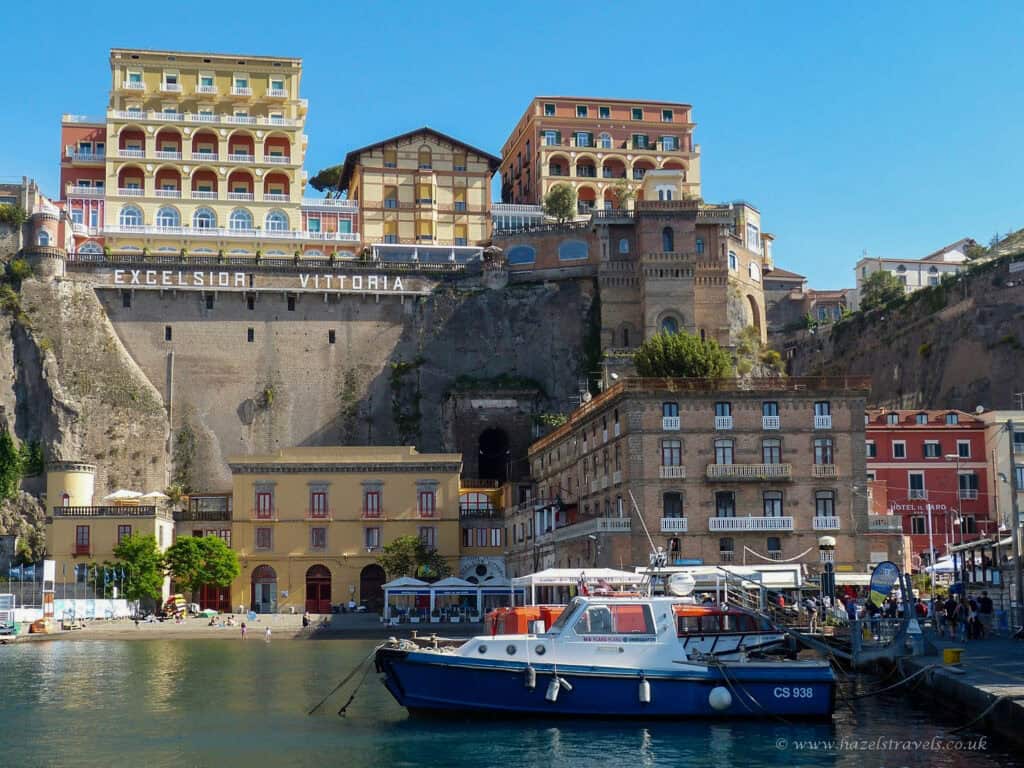 Marina Grande in Sorrento, Italy - colourful waterfront buildings and boats moored in the harbour, with the grand Excelsior Vittoria hotel perched dramatically on the cliff above.