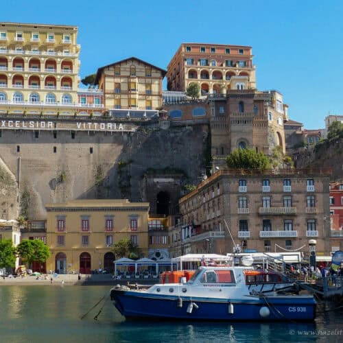 Marina Grande in Sorrento, Italy - colourful waterfront buildings and boats moored in the harbour, with the grand Excelsior Vittoria hotel perched dramatically on the cliff above.