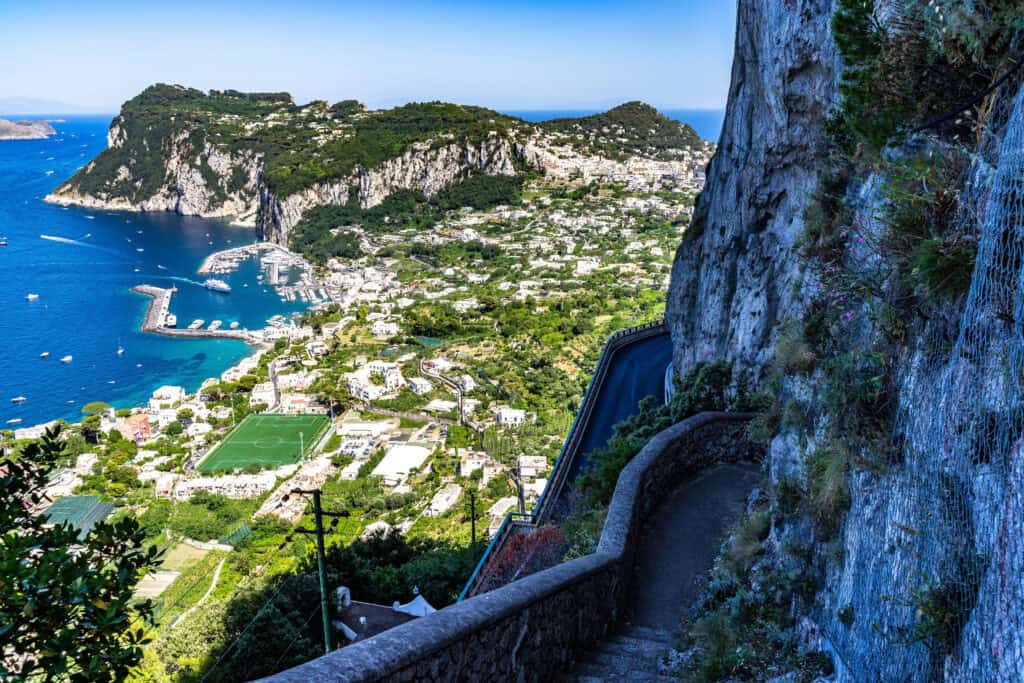 Scenic view from a cliffside path overlooking the island of Capri, Italy, with winding roads, lush greenery, and the sparkling blue waters of the Tyrrhenian Sea dotted with boats below.