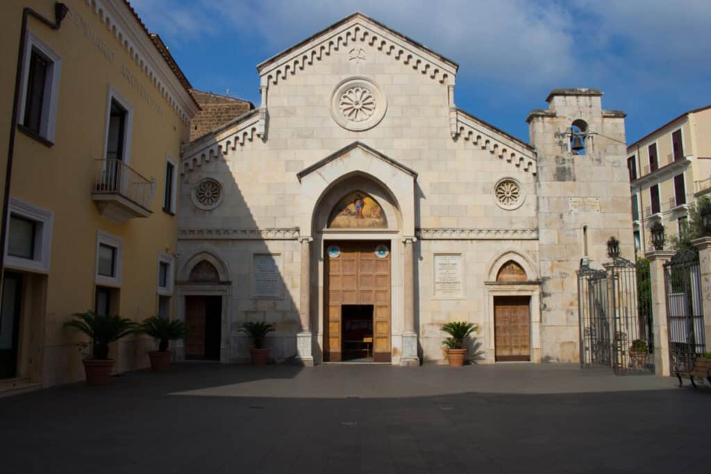 Front façade of Sorrento Cathedral in Italy, featuring Romanesque-style stone architecture, arched doorways, and a small bell tower, surrounded by yellow buildings and potted palms.