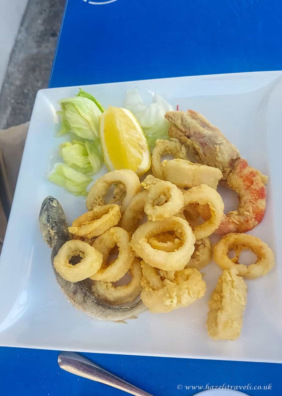 Calamari fritti, La Maddalena - Plate of golden fried calamari with lemon wedges and salad on a blue table.