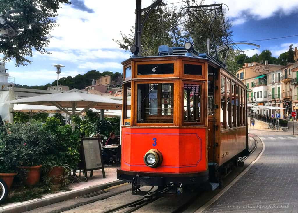 Vintage orange wooden tram in Sóller, Mallorca, travelling along a track through a charming town street lined with cafés, potted plants, and traditional Mediterranean buildings.