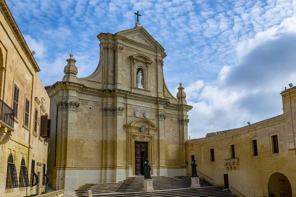 Cathedral of the Assumption in Victoria, Gozo, with statues on the steps and a bright blue sky overhead.
