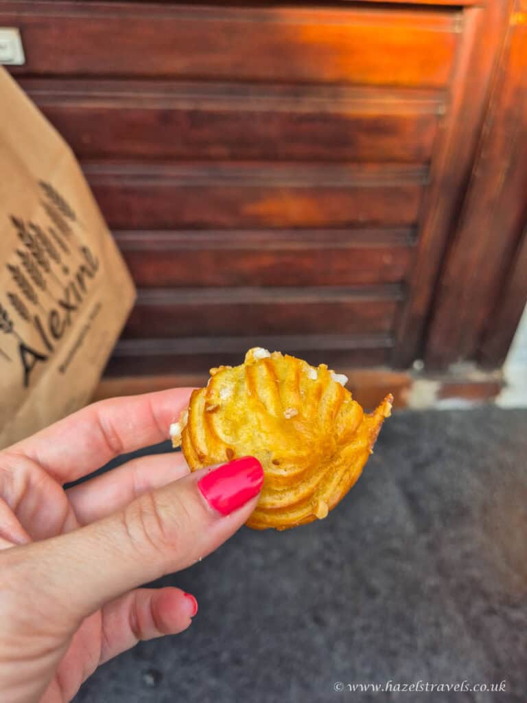 Hand holding a golden chouquette pastry outside a Paris boulangerie, with a paper bag and wooden shopfront in the background.