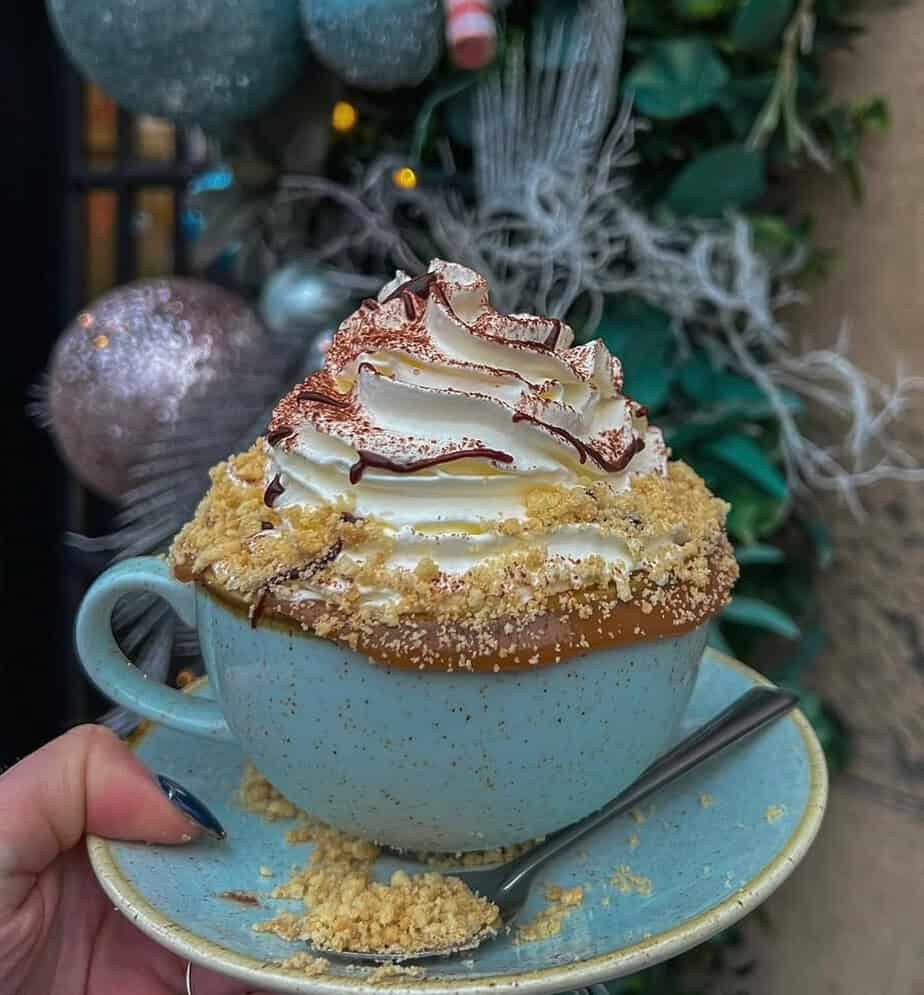 Turquoise mug filled with hot chocolate, topped with white whipped cream, chocolate drizzle, and golden biscuit crumbs, held in front of a festive green and silver Christmas display.