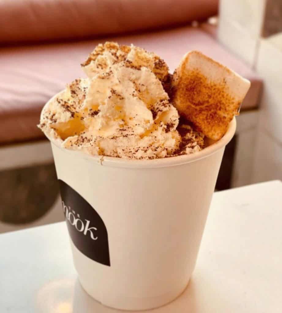 Cup of hot chocolate topped with whipped cream and a toasted marshmallow, served in a cream-coloured takeaway cup.