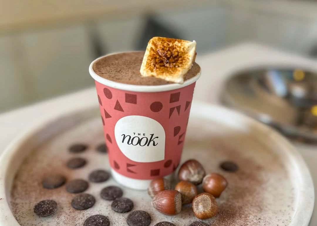 Pink festive takeaway cup of hot chocolate topped with a caramelised toasted marshmallow, surrounded by cocoa powder and chocolate drops.