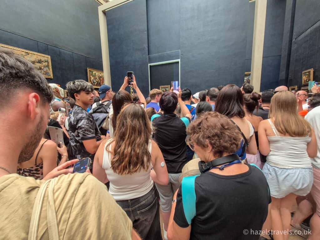 Crowds gathered inside the Louvre Museum in Paris, with visitors holding up phones to photograph the Mona Lisa from a distance.