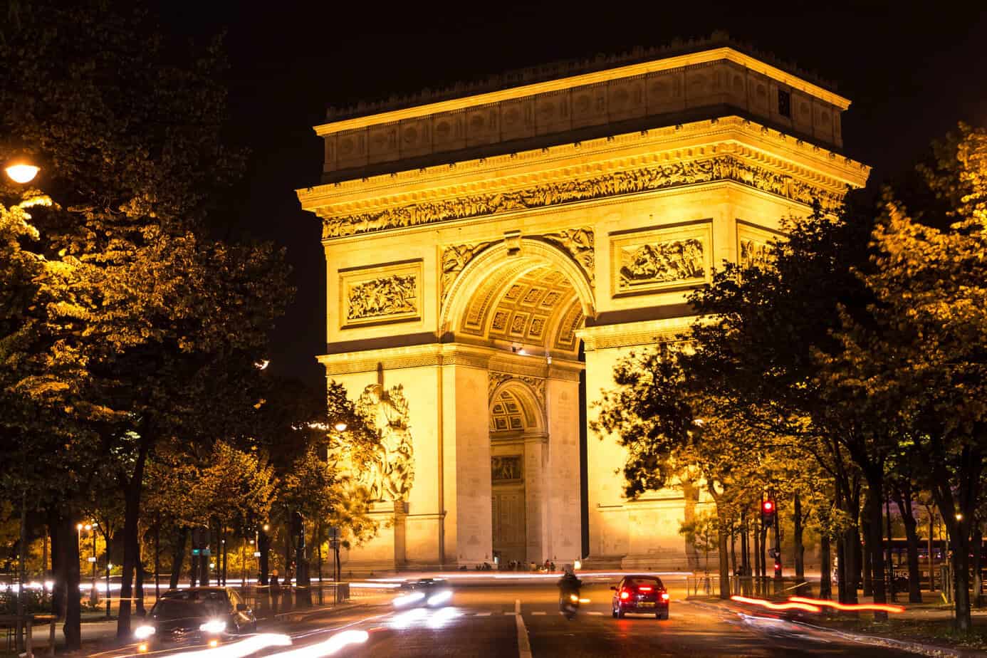 Arc de Triomphe illuminated at night in Paris, glowing golden against a deep blue evening sky.