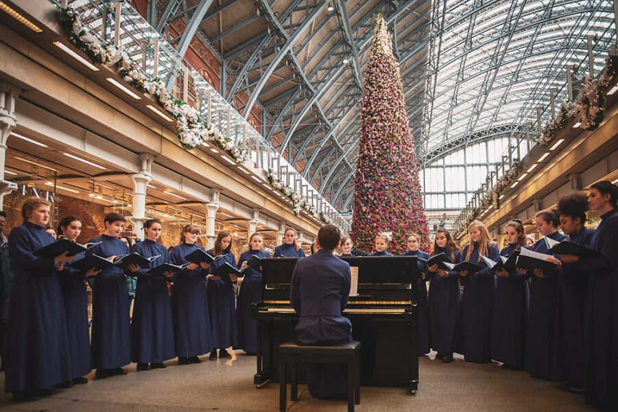 Choir dressed in navy robes singing around a pianist beneath St Pancras Internationalâs towering floral Christmas tree, with garlands and the stationâs blue steel arches overhead.
