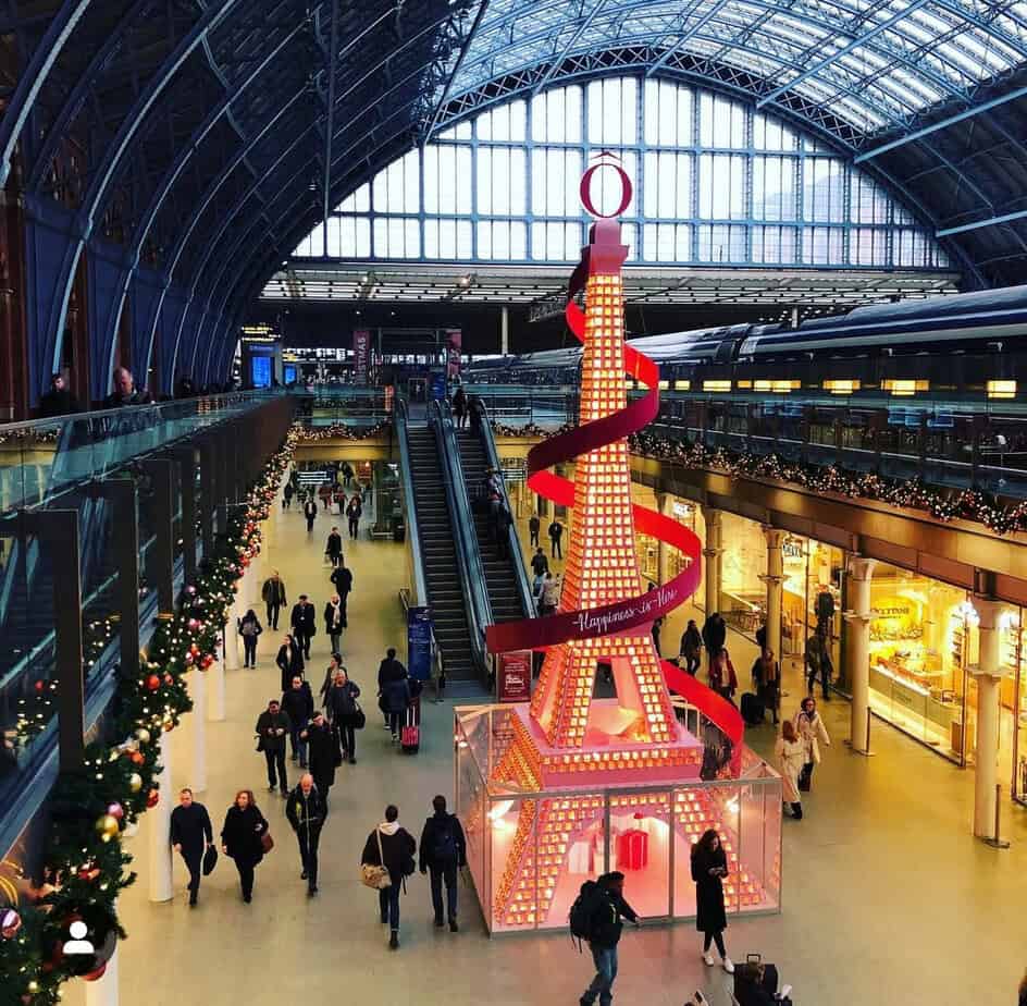 Eiffel Towerâthemed Christmas tree installation at St Pancras International, glowing with warm orange lights and wrapped in a red ribbon, set beneath the stationâs blue steel and glass roof as travellers walk through the concourse.