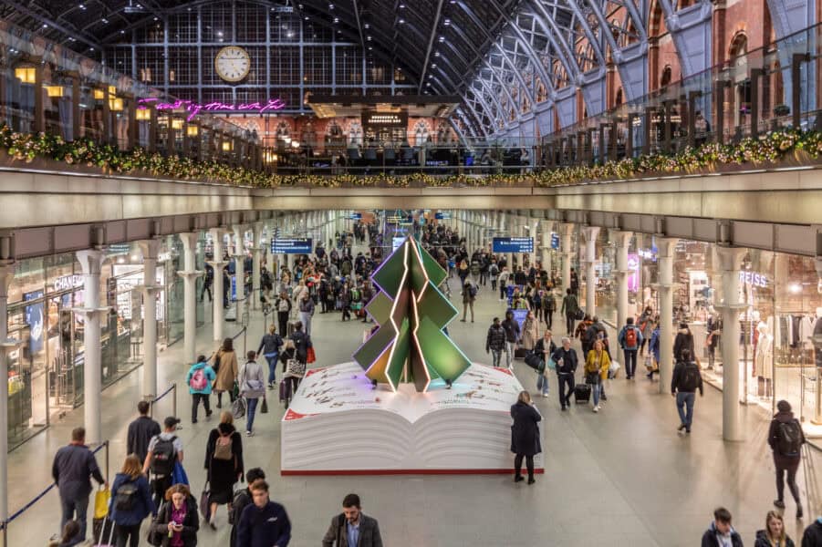 St Pancras Internationalâs storybook-themed Christmas tree featuring a giant open book with a stylised green paper-tree sculpture on top, surrounded by busy travellers and festive garlands beneath the stationâs arched glass roof.