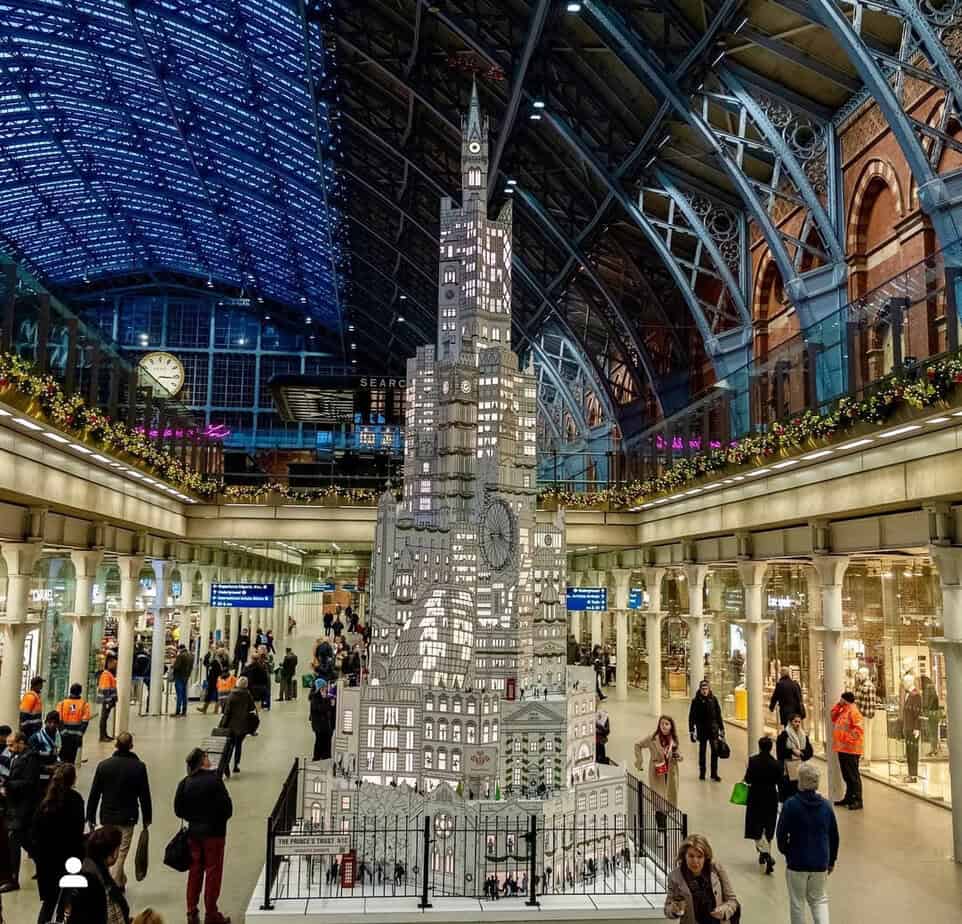 Tall shimmering Christmas tree installation at St Pancras International, decorated in silver and white lights beneath the blue-tinted glass roof.
