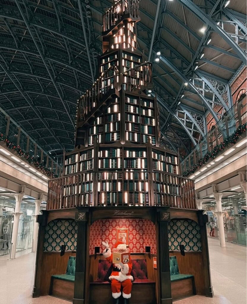 Towering Christmas tree made entirely of stacked books in shades of red, green and gold at St Pancras station, glowing with warm lights.