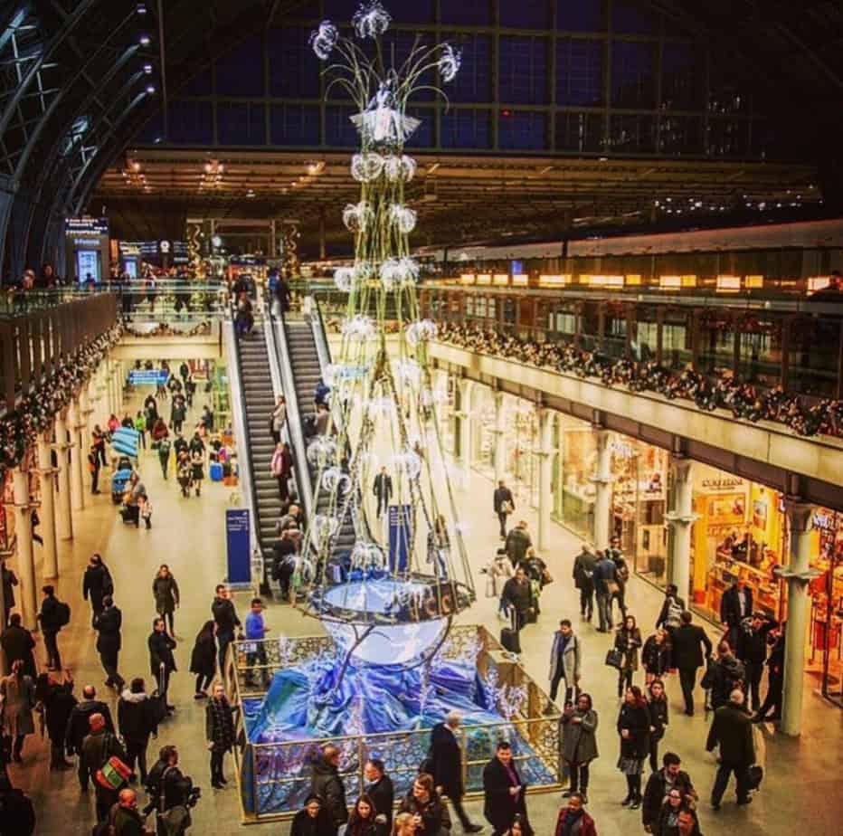 Festive scene at St Pancras with a blue-lit ferris wheel, sparkling white lights and a busy Christmas market under the stationâs high glass ceiling.