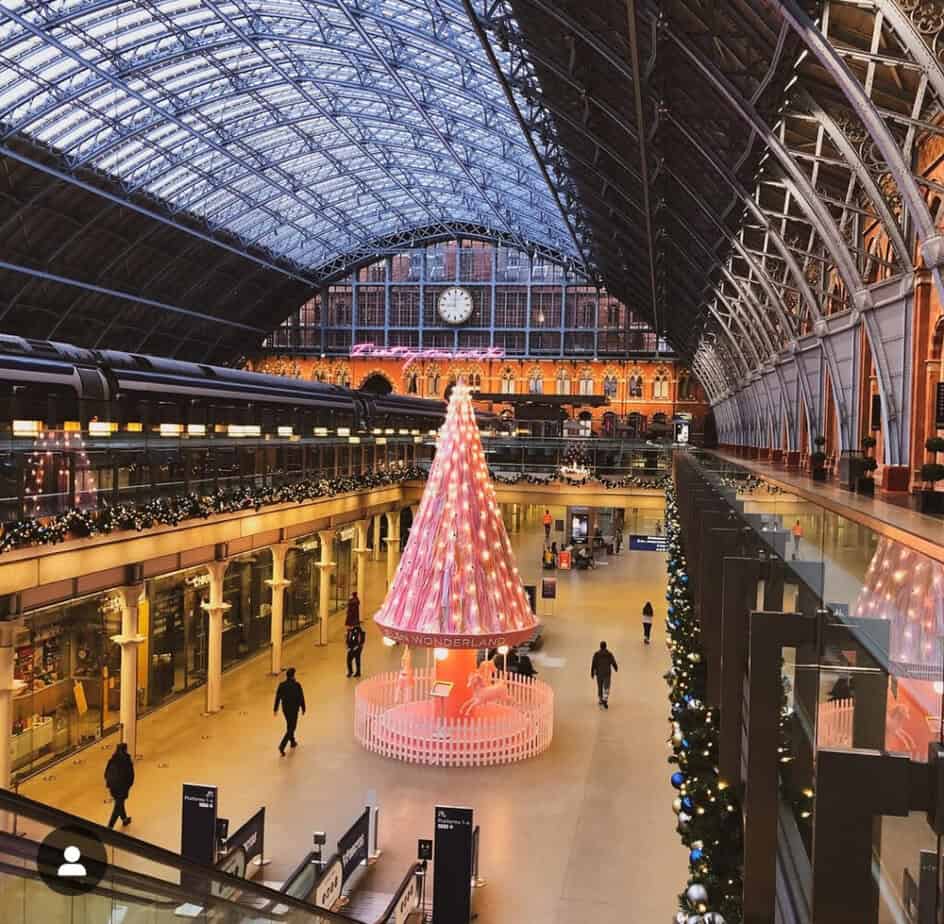 Pink and white illuminated Christmas tree glowing in the centre of St Pancras International, set beneath the stationâs blue-steel arched roof.