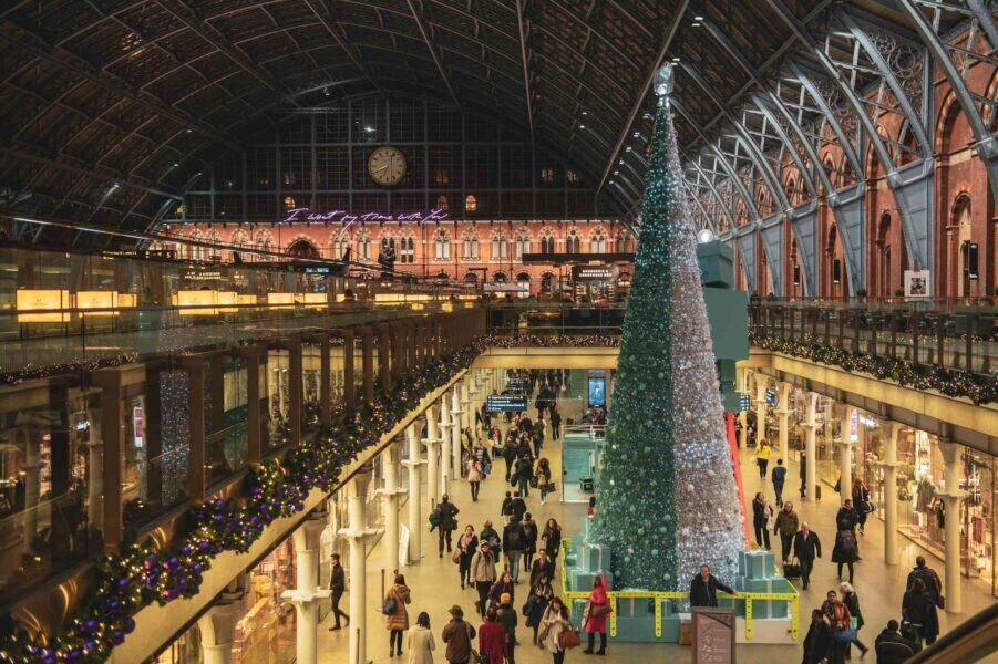 Giant shimmering silver and teal Christmas tree at St Pancras International, surrounded by turquoise gift boxes and festive garlands, with travellers walking beneath the stationâs dramatic arched glass roof and illuminated clock.
