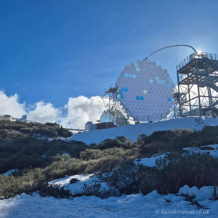 Large telescope dish at Roque de los Muchachos Observatory in La Palma, Canary Islands, surrounded by snow and vegetation under a bright blue sky.