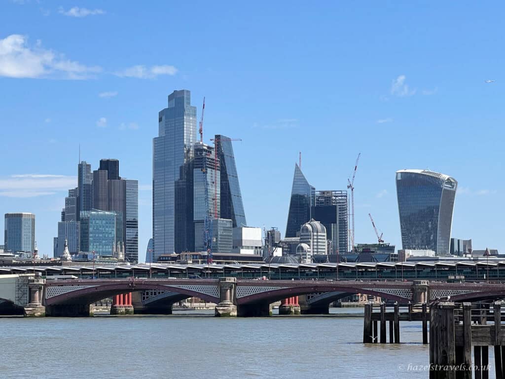 London skyline with modern skyscrapers including the Walkie Talkie and the Cheesegrater, viewed from across the River Thames under a clear blue sky.